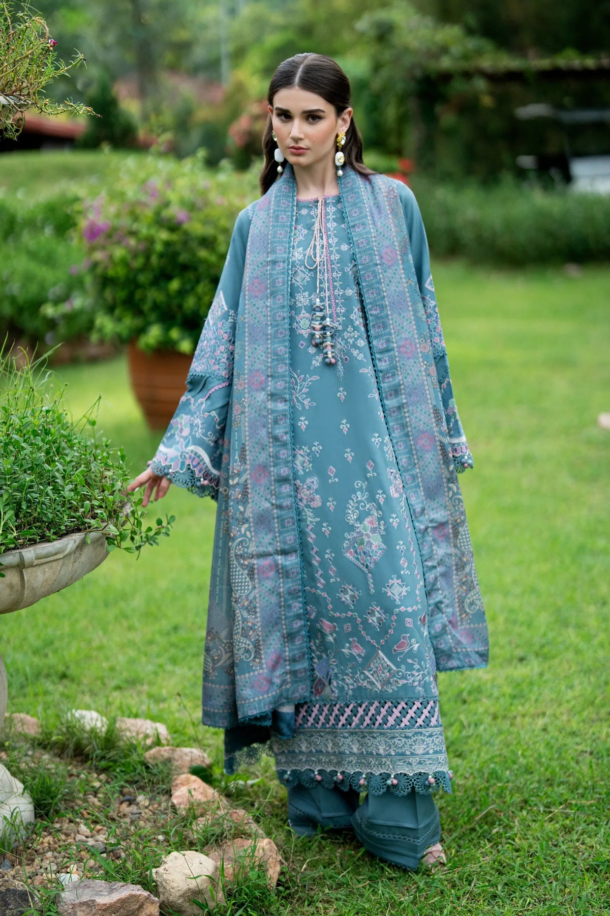 A woman stands on a grassy garden lawn wearing a long teal-blue printed traditional outfit with matching pants. The scene includes a stone planter and blurred greenery in the background.