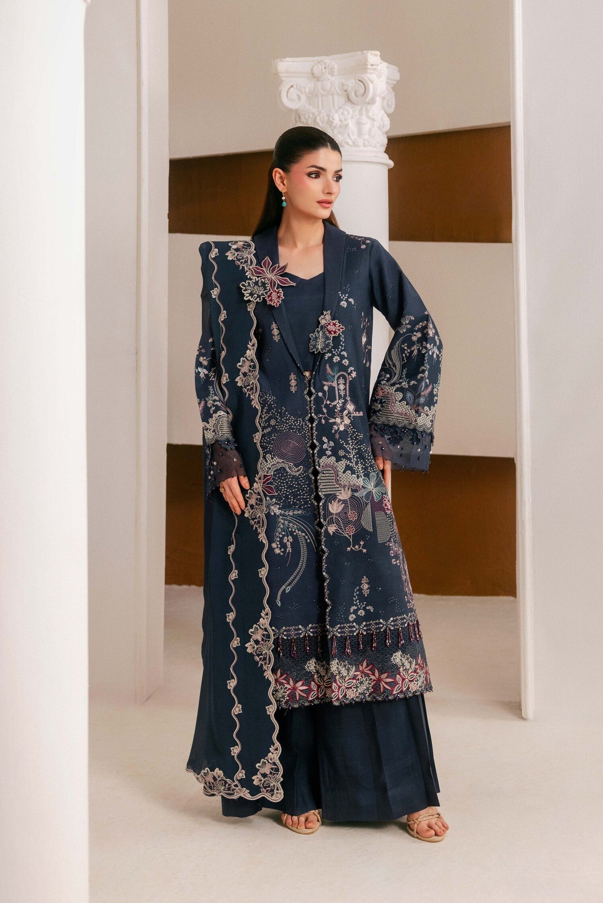 A woman models a dark navy blue traditional outfit with intricate floral embroidery and a matching dupatta, standing in a modern architectural setting with white columns and brown accent walls.